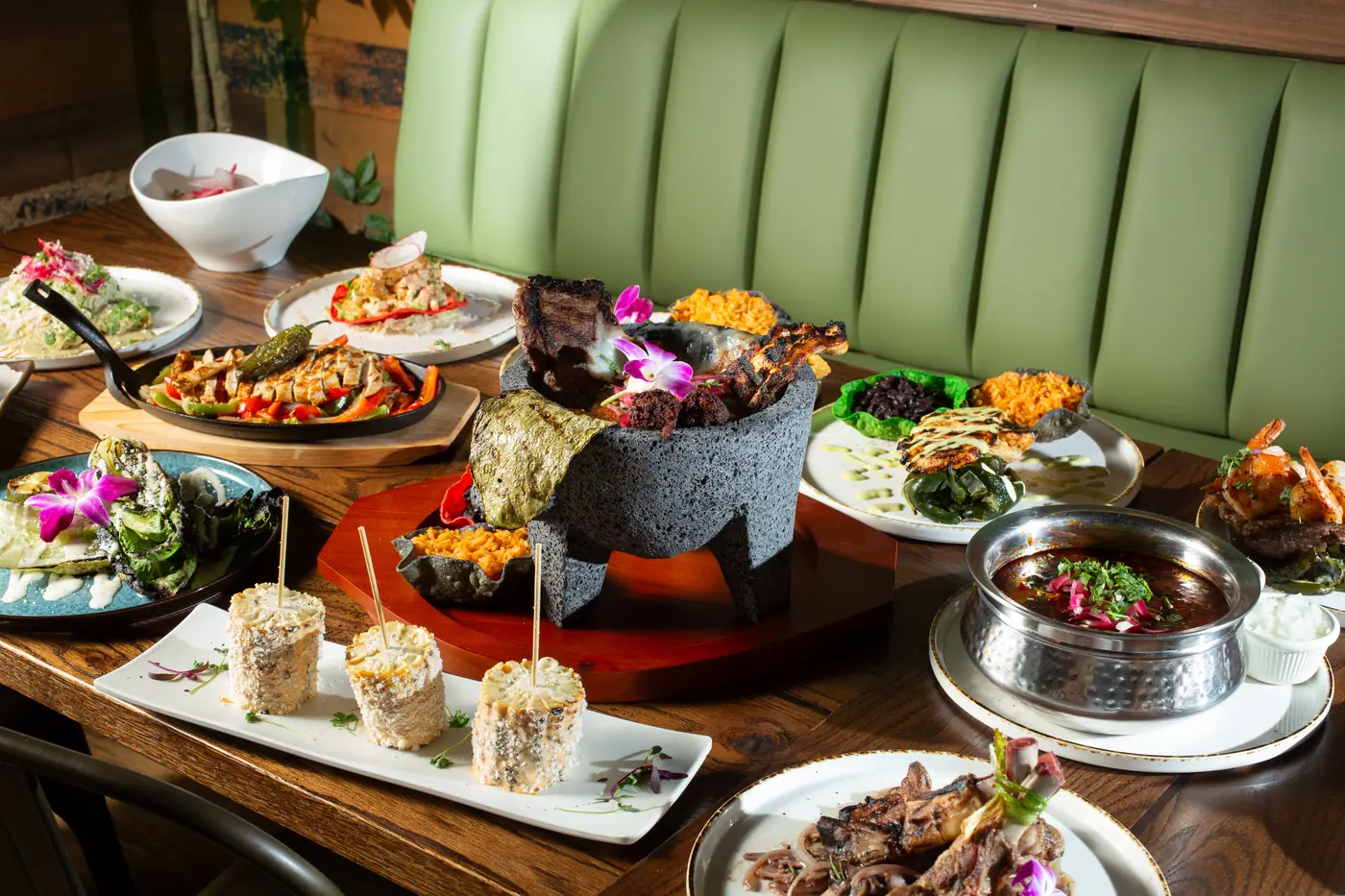 A spread of OMA dishes on a wooden table: a stone molcajete of grilled meats, sizzling chicken fajitas, elote, grilled romaine, shrimp and a bowl of birria